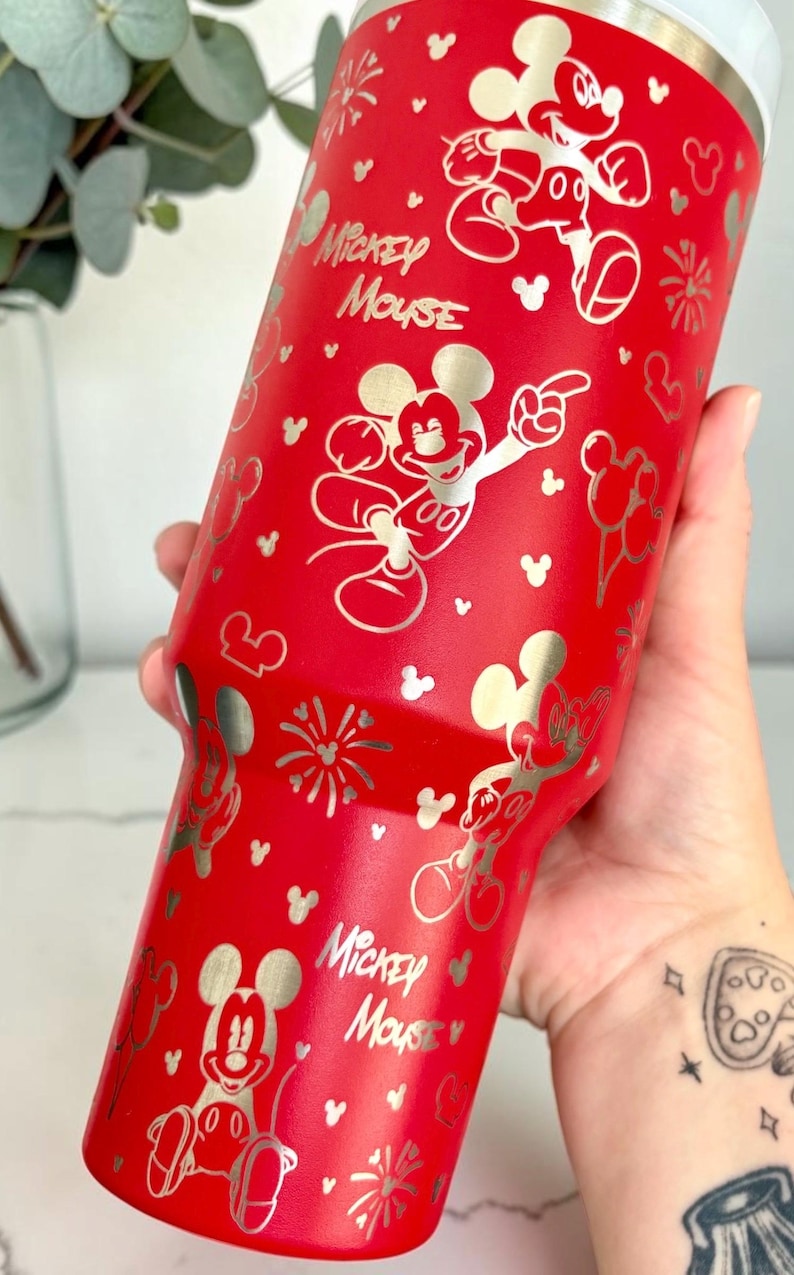 May include: A red tumbler featuring a silver Mickey Mouse design. The tumbler has a silver rim and is covered in Mickey Mouse illustrations, fireworks, and the words "Mickey Mouse".