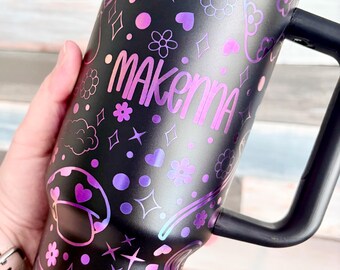 May include: Black travel mug with a handle, decorated with a pattern of flowers, hearts, stars, and mushrooms in shades of purple and pink. The name "Makenna" is printed in a gradient font. The mug has a black lid and handle.