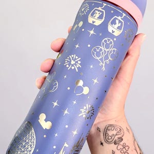 Op de afbeelding: Een paarse waterfles met een goudkleurig patroon van Disney-iconen zoals kastelen, gondels en ballonnen. De dop is lichtpaars met een donkerpaarse sluiting waarop de tekst Lift to Unlock staat gedrukt in een wit kader.
