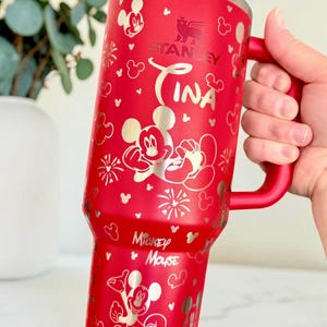 May include: Red Stanley tumbler with a handle, featuring Mickey Mouse and the name "Tina" in silver. The tumbler has a white lid and is decorated with Mickey Mouse silhouettes and floral designs.