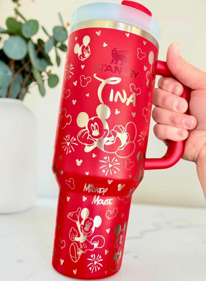 May include: Red Stanley tumbler with a handle, featuring Mickey Mouse and the name "Tina" in silver. The tumbler has a white lid and is decorated with Mickey Mouse silhouettes and floral designs.