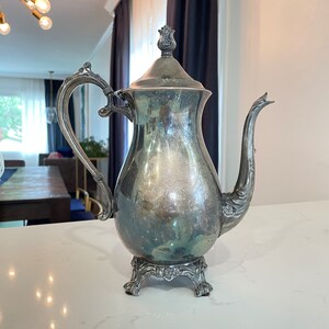May include: A silver teapot with a decorative handle and a floral design on the lid. The teapot has a pearlescent finish and sits on three ornate feet.