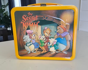 May include: Yellow metal lunchbox featuring a cartoon image of characters from the movie "The Secret of NIMH". The characters are mice and a rat, and they are standing in front of a cave. The lunchbox has a black handle.