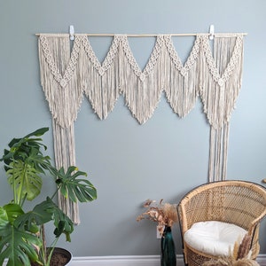May include: A macrame wall hanging with a chevron pattern in a cream colour. The hanging is made of cotton cord and is attached to a wooden dowel rod. The hanging is displayed on a light blue wall with a potted plant and a wicker chair in the foreground.