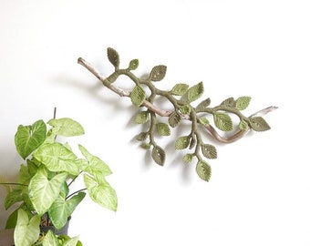May include: A macrame wall hanging in the shape of a branch with green leaves. The branch is made of natural wood and the leaves are made of yarn. The wall hanging is mounted on a white wall.
