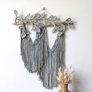 May include: A gray macrame wall hanging with a branch and leaf design. The wall decor features long fringe and is suspended from a wooden dowel. A vase with dried flowers and a potted plant are on a wooden table.