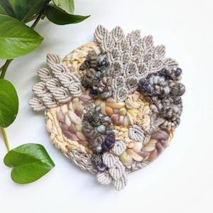 May include: A circular wall hanging made with macrame and woven yarn in shades of beige, brown, and purple. The piece features a variety of textures and patterns, including a central circle of woven yarn and a border of macrame leaves.