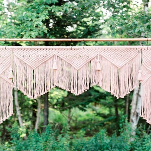 May include: A pink macrame wedding arch with tassels hangs from a copper pipe frame. The arch is in front of a green leafy background.