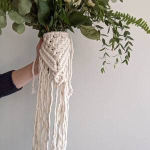 Op de afbeelding: Een witte macramé bloempot met een lange franje. De pot is gevuld met een boeket witte rozen en groen.