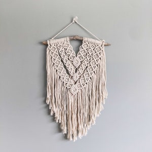 May include: A white macrame wall hanging with a wooden dowel at the top. The macrame is in a V-shape with fringe hanging down the sides.