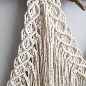 Pode incluir: Uma tape&ccedil;aria de macram&eacute; branca com um padr&atilde;o geom&eacute;trico e franjas. A tape&ccedil;aria est&aacute; presa a um peda&ccedil;o de madeira flutuante.