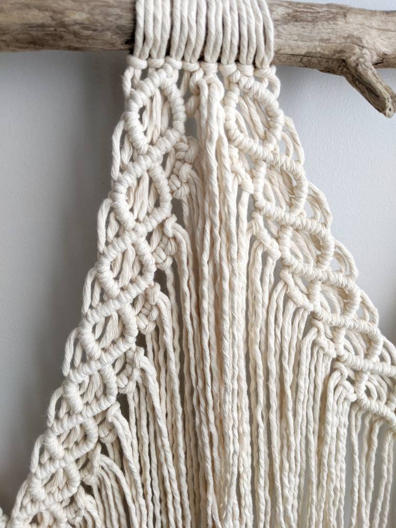 Pode incluir: Uma tape&ccedil;aria de macram&eacute; branca com um padr&atilde;o geom&eacute;trico e franjas. A tape&ccedil;aria est&aacute; presa a um peda&ccedil;o de madeira flutuante.