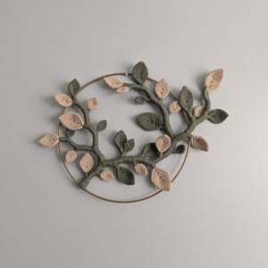Peut inclure: Décoration murale en macramé avec un anneau en métal bronze formant un cercle. Le design présente une structure en forme de branche avec des feuilles vertes et beiges. Les feuilles sont nouées, créant une esthétique texturée et naturelle. Idéal pour la décoration intérieure.