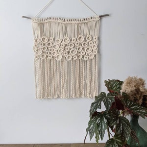 Könnte beinhalten: Cremefarbener Makramee-Wandbehang mit geometrischem Blumendesign. Der Makramee wird an einer natürlichen Holzdübel und einer cremefarbenen Kordel aufgehängt. Der Wandbehang wird vor einer weißen Wand ausgestellt.