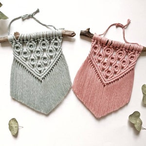 Könnte beinhalten: Zwei Makramee-Wandbehänge mit geometrischem Muster, einer in Hellblau und einer in Rosa, jeweils mit einem Naturholzast oben.
