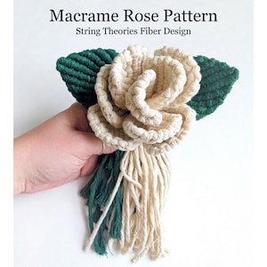 以下が含まれることがあります： クリーム色のマクラメのバラで、緑の葉とフリンジが付いています。「Macrame Rose Pattern」と「String Theories Fiber Design」の文字が見えます。手作りのマクラメの花は装飾品です。