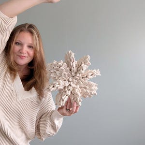 May include: A person is holding a large, white, crocheted snowflake ornament. The snowflake is made of yarn and has a delicate, lacy design.