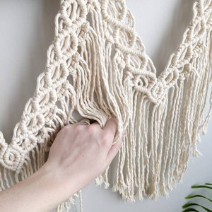 Pode incluir: Uma tape&ccedil;aria de macram&eacute; de parede com um cord&atilde;o de algod&atilde;o de cor creme, com um padr&atilde;o geom&eacute;trico e detalhes de franjas. A tape&ccedil;aria est&aacute; presa a um ramo de madeira natural.