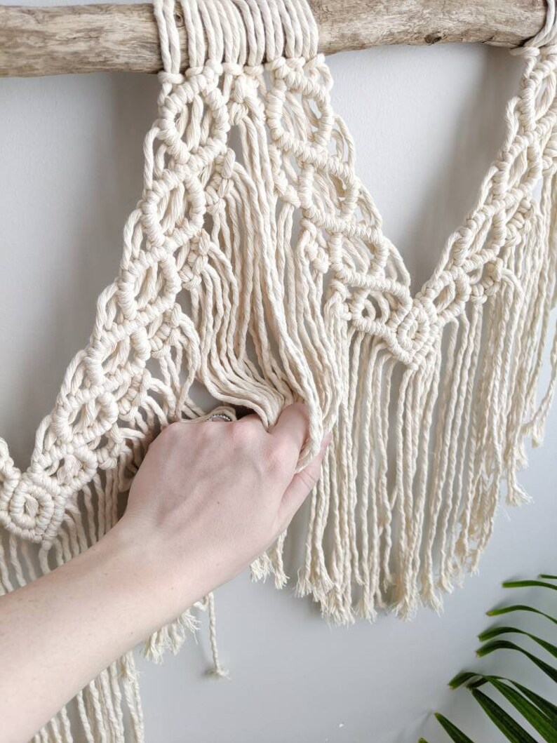 Pode incluir: Uma tape&ccedil;aria de macram&eacute; de parede com um cord&atilde;o de algod&atilde;o de cor creme, com um padr&atilde;o geom&eacute;trico e detalhes de franjas. A tape&ccedil;aria est&aacute; presa a um ramo de madeira natural.