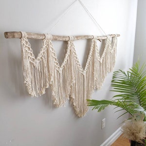 Pode incluir: Uma tape&ccedil;aria de macram&eacute; de parede com um ramo de madeira natural e um cord&atilde;o de algod&atilde;o branco. A tape&ccedil;aria apresenta uma s&eacute;rie de formas geom&eacute;tricas e franjas.