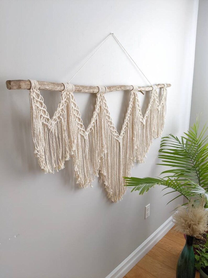 Pode incluir: Uma tape&ccedil;aria de macram&eacute; de parede com um ramo de madeira natural e um cord&atilde;o de algod&atilde;o branco. A tape&ccedil;aria apresenta uma s&eacute;rie de formas geom&eacute;tricas e franjas.