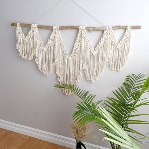 Pode incluir: Uma tape&ccedil;aria de macram&eacute; de parede com um ramo de madeira natural, apresentando um padr&atilde;o geom&eacute;trico com um cord&atilde;o de algod&atilde;o branco. A tape&ccedil;aria &eacute; feita de m&uacute;ltiplos tri&acirc;ngulos com detalhes de franjas.
