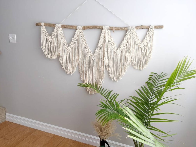 Pode incluir: Uma tape&ccedil;aria de macram&eacute; de parede com um ramo de madeira natural, apresentando um padr&atilde;o geom&eacute;trico com um cord&atilde;o de algod&atilde;o branco. A tape&ccedil;aria &eacute; feita de m&uacute;ltiplos tri&acirc;ngulos com detalhes de franjas.