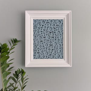 Könnte beinhalten: Ein gerahmtes Kunstwerk mit einem blauen Makramee-Design, das ein sich wiederholendes kreisförmiges Muster zeigt. Das Kunstwerk befindet sich in einem weißen Holzrahmen und hängt an einer grauen Wand. Grüne Pflanzenblätter sind unten links sichtbar.