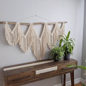 Pode incluir: Uma tape&ccedil;aria de macram&eacute; de parede com um ramo de madeira natural, apresentando um design geom&eacute;trico de cinco pontos. A tape&ccedil;aria &eacute; de cor creme e &eacute; exibida em uma parede branca acima de uma mesa consola de madeira com duas gavetas. Duas plantas em vaso est&atilde;o sobre a mesa.