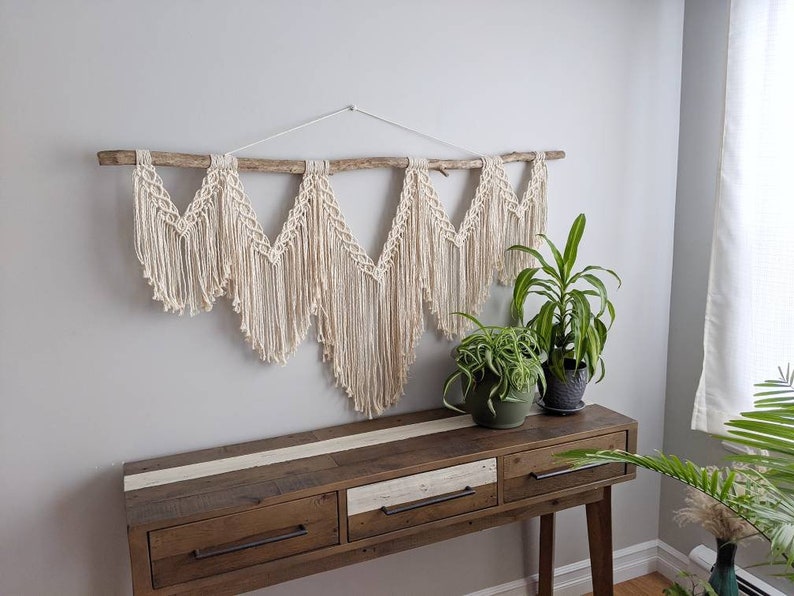Pode incluir: Uma tape&ccedil;aria de macram&eacute; de parede com um ramo de madeira natural, apresentando um design geom&eacute;trico de cinco pontos. A tape&ccedil;aria &eacute; de cor creme e &eacute; exibida em uma parede branca acima de uma mesa consola de madeira com duas gavetas. Duas plantas em vaso est&atilde;o sobre a mesa.