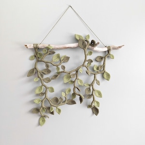 May include: A handcrafted wall hanging featuring a macrame design of leaves and branches in shades of green and brown. The artwork is suspended from a natural wooden branch with a small bird accent, creating a nature-inspired decorative piece.