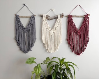 Pode incluir: Três tapeçarias de macramé de parede com um ramo de madeira no topo. As tapeçarias são de cor cinza, branca e bordô.