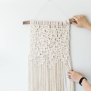 May include: A white macrame wall hanging with a wooden dowel rod at the top and a fringe at the bottom.
