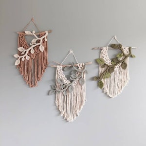 May include: Three macrame wall hangings in shades of pink, cream, and green, each featuring a wooden dowel and a decorative branch with leaves. The wall art is displayed against a gray wall, showcasing a bohemian style.