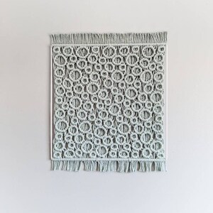 Könnte beinhalten: Ein Wandbehang aus Makramee in hellgrüner Farbe mit einem kreisförmigen Muster. Der Behang hat einen Fransenrand.