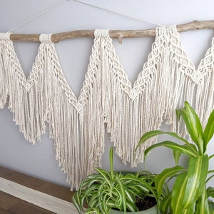 Pode incluir: Uma tape&ccedil;aria de macram&eacute; de parede com um ramo de madeira natural, apresentando um padr&atilde;o geom&eacute;trico com franjas. A tape&ccedil;aria &eacute; feita de cord&atilde;o de algod&atilde;o branco e &eacute; exibida contra uma parede cinza claro.