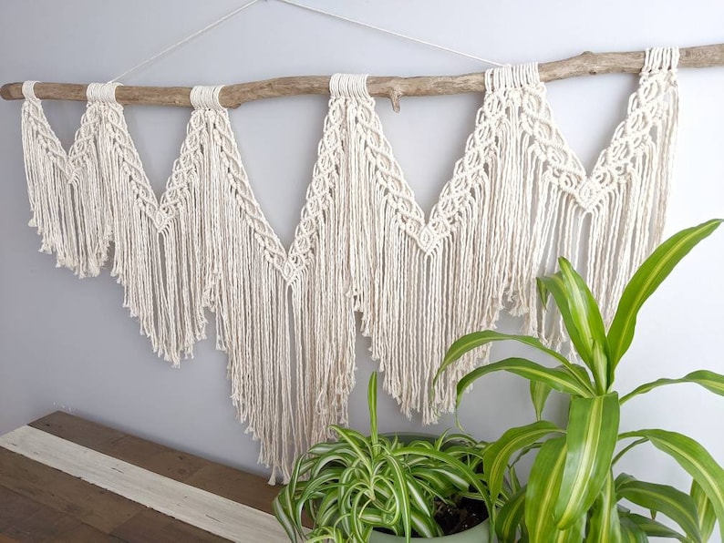 Pode incluir: Uma tape&ccedil;aria de macram&eacute; de parede com um ramo de madeira natural, apresentando um padr&atilde;o geom&eacute;trico com franjas. A tape&ccedil;aria &eacute; feita de cord&atilde;o de algod&atilde;o branco e &eacute; exibida contra uma parede cinza claro.