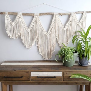 Pode incluir: Uma tape&ccedil;aria de macram&eacute; branca com um ramo de madeira natural, pendurada acima de uma mesa de consola de madeira com tr&ecirc;s gavetas. Duas plantas em vasos est&atilde;o sobre a mesa.