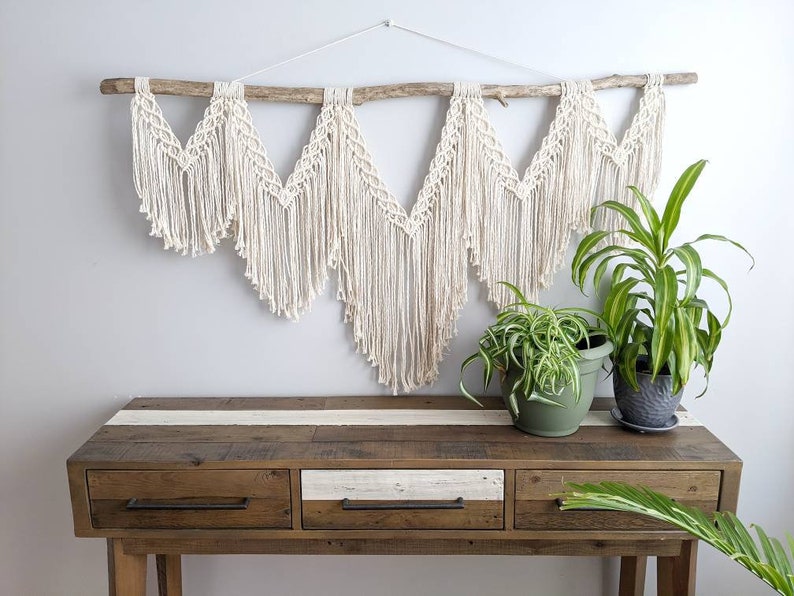 Pode incluir: Uma tape&ccedil;aria de macram&eacute; branca com um ramo de madeira natural, pendurada acima de uma mesa de consola de madeira com tr&ecirc;s gavetas. Duas plantas em vasos est&atilde;o sobre a mesa.