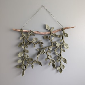 May include: A macrame wall hanging featuring a branch with green leaves. The branch is made of driftwood and the leaves are made of knotted string.