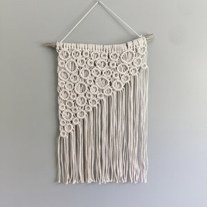 Könnte beinhalten: Cremefarbener Makramee-Wandbehang. Das Design zeigt eine Reihe miteinander verbundener Kreise oben links, die in lange, vertikale Fransen übergehen. Es ist an einem natürlichen Holzdübel und einer weißen Schnur aufgehängt.