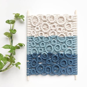 Könnte beinhalten: Ein Wandbehang aus Makramee in den Farben Blau, Hellblau und Weiß. Das Makramee ist mit einem Knotenmuster gearbeitet, das an Kreise erinnert. Der Wandbehang ist an zwei Holzstäben befestigt.