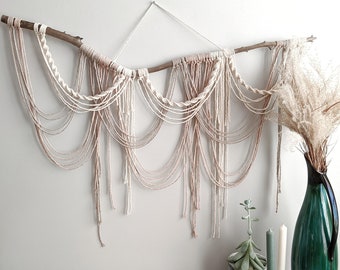 May include: A macrame wall hanging with a natural branch and cream and blush colored yarn. The hanging is displayed on a white wall with a green vase and a succulent plant on a wooden table.