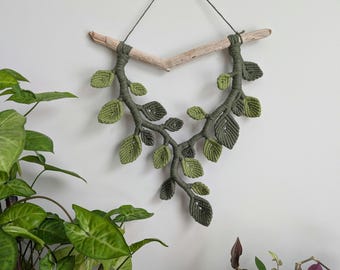 May include: A macrame wall hanging featuring a branch with green leaves. The branch is made of driftwood and the leaves are made of knotted string.