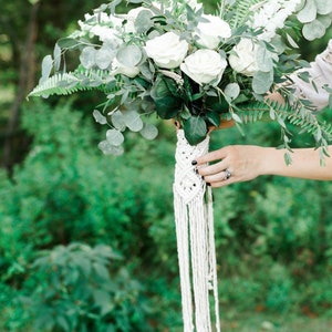 Peut inclure: Un bouquet de mariée blanc avec des roses blanches, de la verdure et des fleurs blanches. Le bouquet est attaché avec une poignée en macramé blanche.