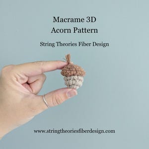 May include: A small, handmade macrame acorn in shades of brown and beige is held between fingers. The acorn has a textured appearance, with the top in a darker brown and the body in a lighter beige. Text on the image reads "Macrame 3D Acorn Pattern" and "String Theories Fiber Design."