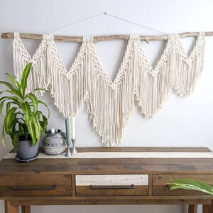 Pode incluir: Uma tape&ccedil;aria de macram&eacute; branca com um ramo de madeira no topo. A tape&ccedil;aria &eacute; feita de uma s&eacute;rie de tri&acirc;ngulos com franjas. A tape&ccedil;aria &eacute; exibida acima de uma mesa consola de madeira com tr&ecirc;s gavetas.