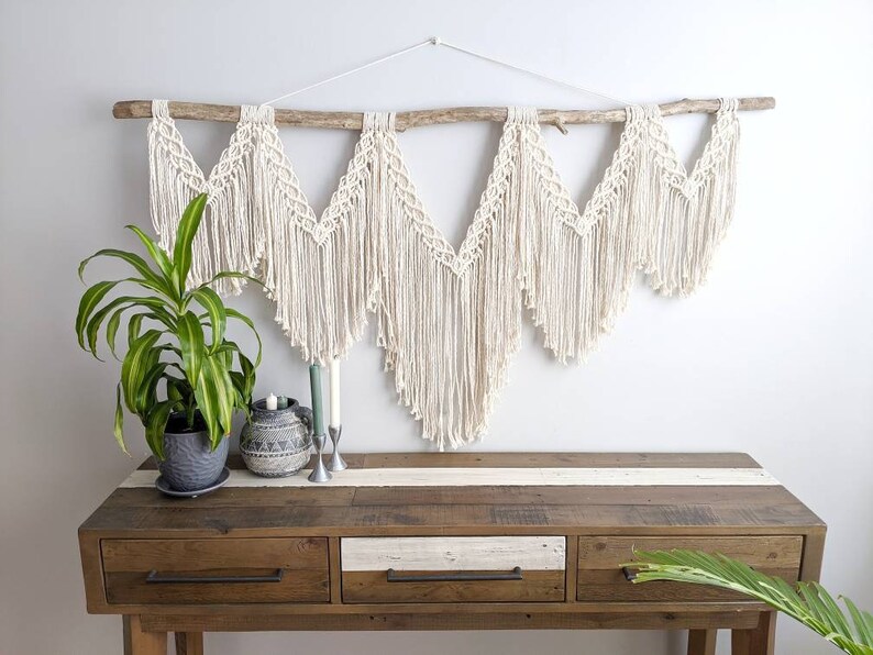 Pode incluir: Uma tape&ccedil;aria de macram&eacute; branca com um ramo de madeira no topo. A tape&ccedil;aria &eacute; feita de uma s&eacute;rie de tri&acirc;ngulos com franjas. A tape&ccedil;aria &eacute; exibida acima de uma mesa consola de madeira com tr&ecirc;s gavetas.