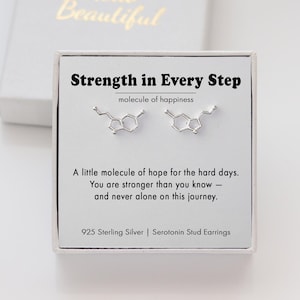 May include: Silver serotonin stud earrings shaped like a molecule, presented on a card with the text "Strength in Every Step" and "molecule of happiness." The card is in a white box with gold lettering that says "Hello Beautiful."