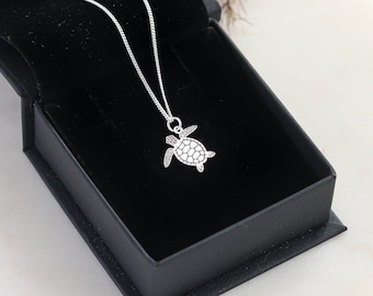 Può includere: Una collana con pendente a forma di tartaruga argentata esposta in un cofanetto nero aperto. Il pendente a forma di tartaruga presenta un design dettagliato del guscio ed è sospeso a una catenina sottile. L'interno della scatola è in velluto nero.